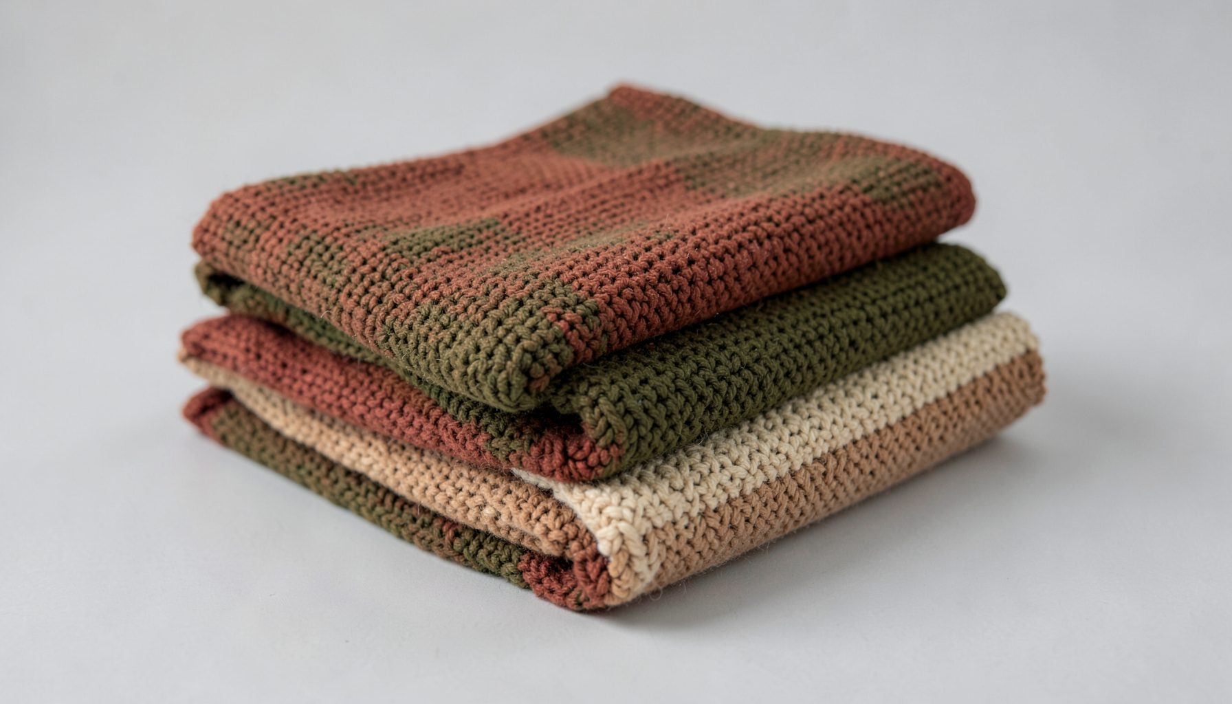 A folded stack of knitted scarves in muted earth tones — rust, olive green, and warm beige — resting on a light grey surface